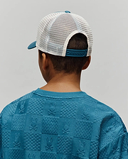 Teal crewneck sweatshirt with a relaxed fit, featuring an all-over embossed checkerboard pattern of Psycho Bunny skull logos and textured squares, paired with a Kids Adamo Trucker Cap, teal front and brim with white mesh back.
