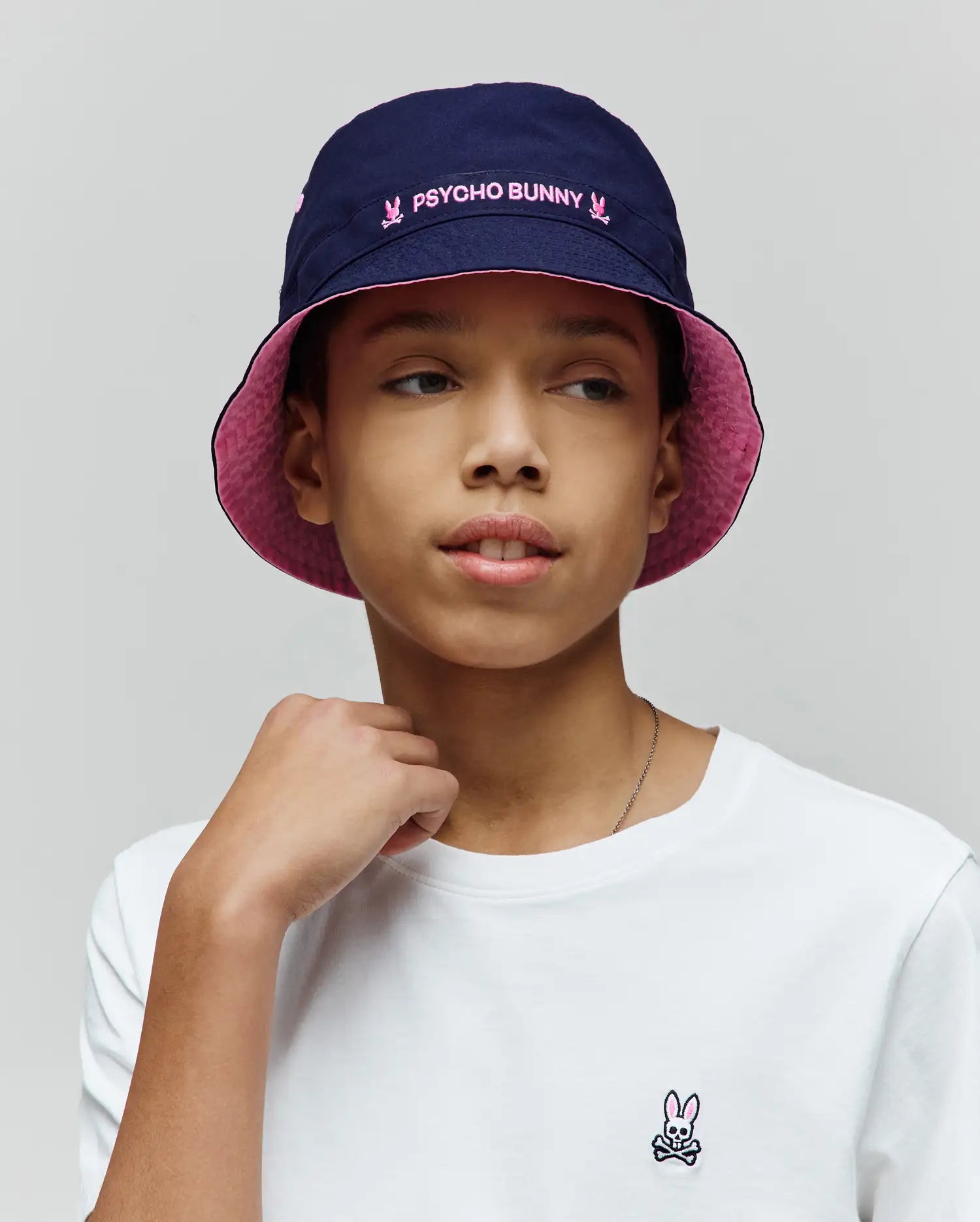Kids Luke Reversible Bucket Hat in navy with pink Psycho Bunny embroidery and a white jersey t-shirt with a small embroidered Psycho Bunny skull logo on the left chest.