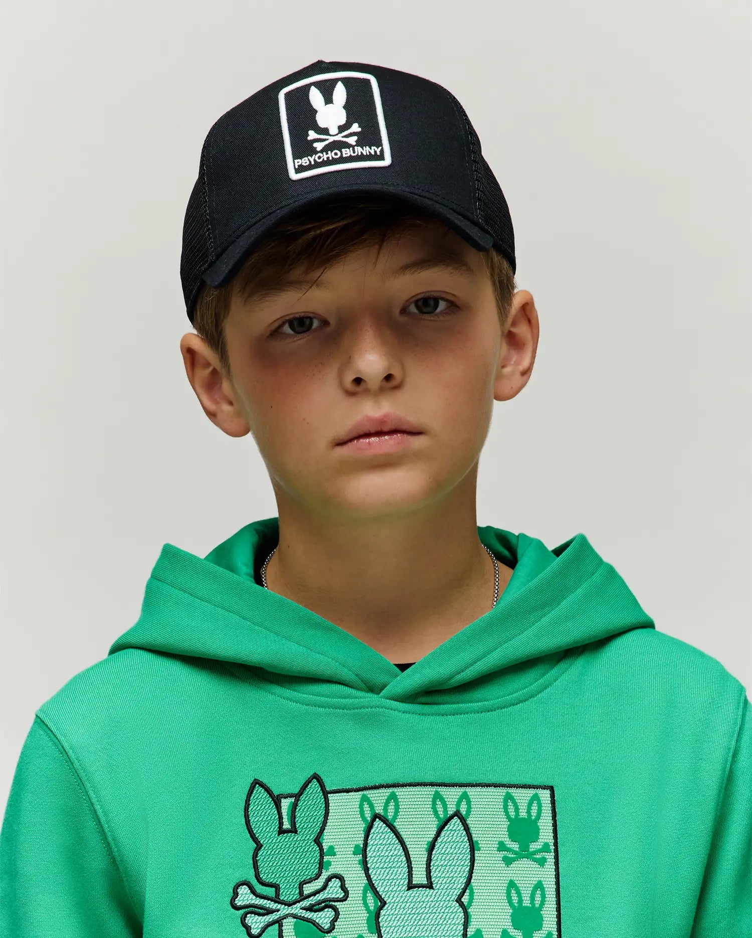 Kids Adamo Trucker Cap, black with white Psycho Bunny logo patch, paired with a bright green jersey knit hoodie featuring a large chest graphic of multiple Psycho Bunny skull logos.