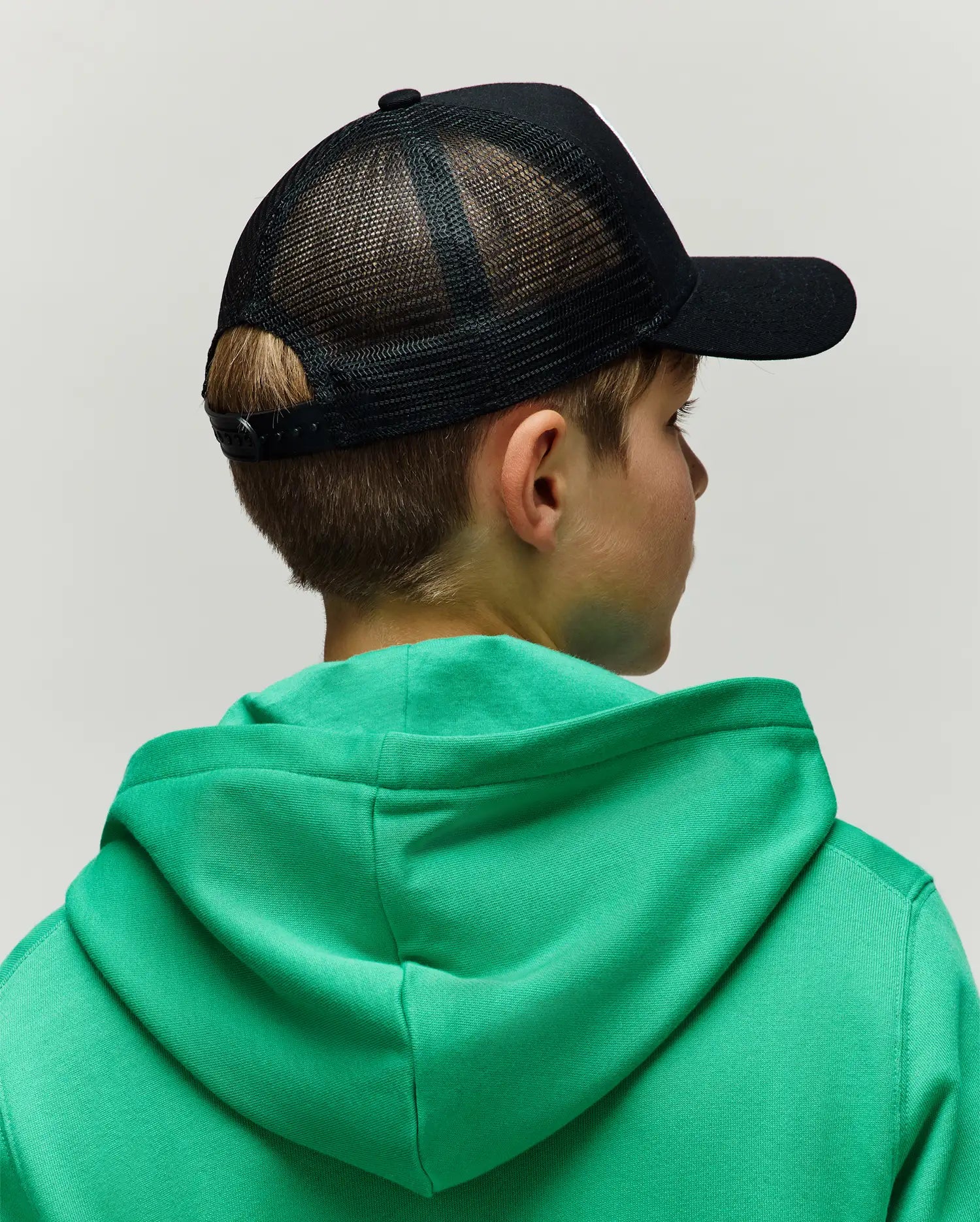 Black KIDS ADAMO TRUCKER CAP with a breathable mesh back and adjustable snap closure, worn with a vibrant green classic fit hooded sweatshirt in a soft, comfortable jersey fabric.