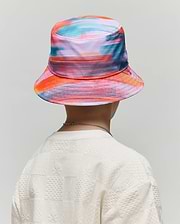 Kids Raphael Bucket Hat in vibrant multi-color abstract print paired with an off-white relaxed fit textured knit t-shirt featuring an all-over checkerboard Psycho Bunny skull logo pattern.