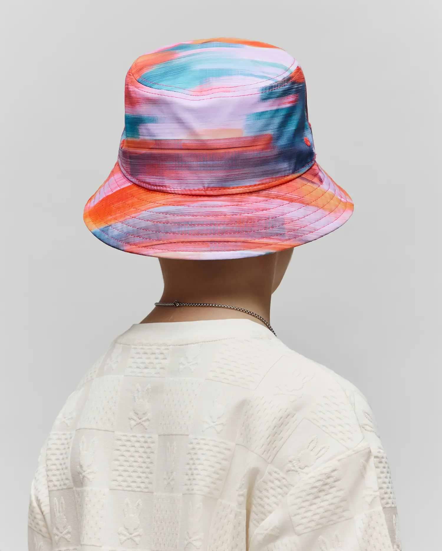 Kids Raphael Bucket Hat in vibrant multi-color abstract print paired with an off-white relaxed fit textured knit t-shirt featuring an all-over checkerboard Psycho Bunny skull logo pattern.