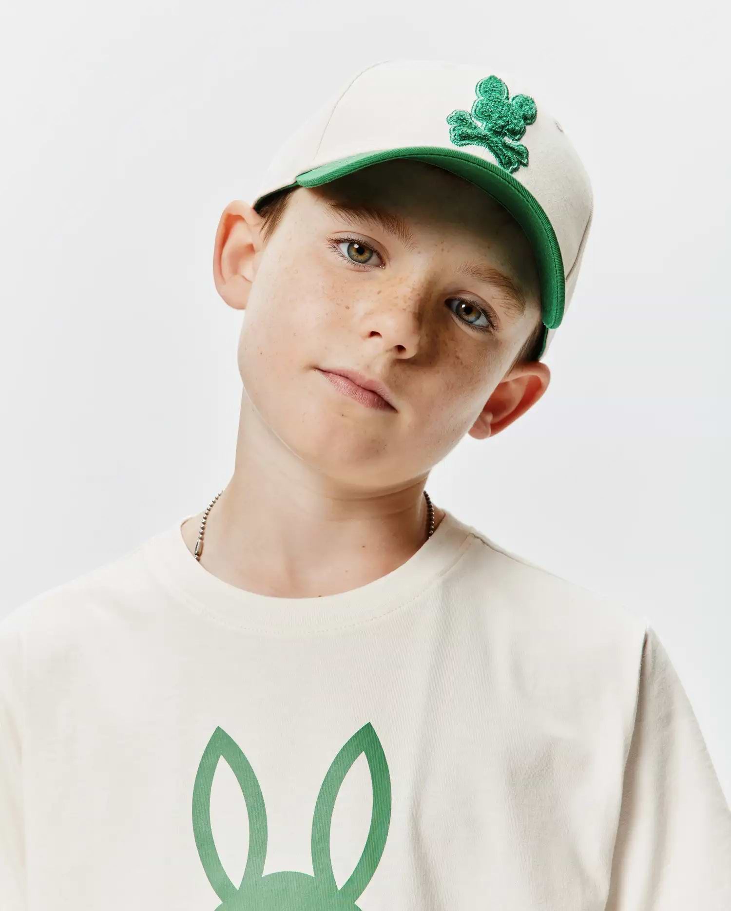 Beige KIDS CRAIG BASEBALL CAP with a green brim and embroidered Psycho Bunny skull logo, complementing an off-white jersey t-shirt featuring a large green bunny ears graphic on the chest.
