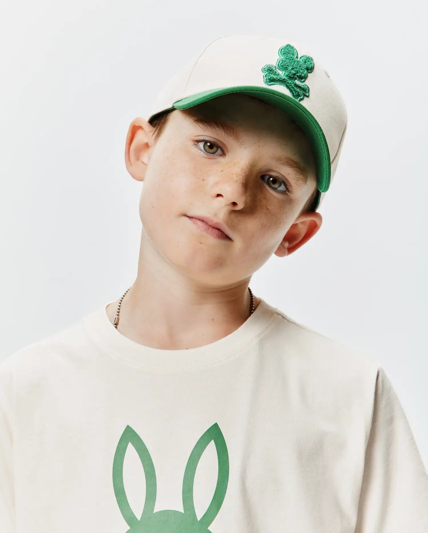 Beige KIDS CRAIG BASEBALL CAP with a green brim and embroidered Psycho Bunny skull logo, complementing an off-white jersey t-shirt featuring a large green bunny ears graphic on the chest.