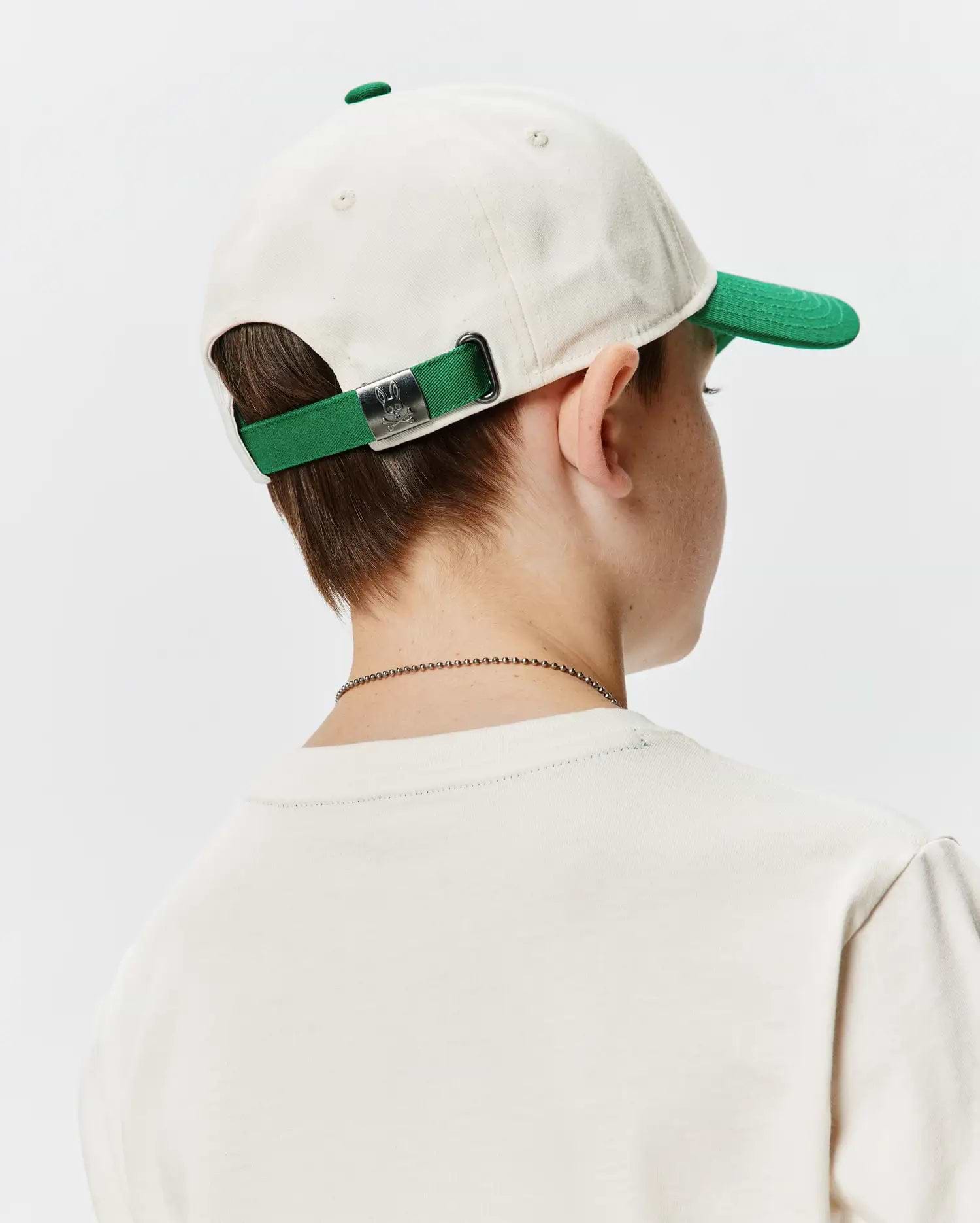 Kids Craig Baseball Cap, an off-white cotton twill cap with a green brim and matching adjustable strap, featuring a silver Psycho Bunny skull logo buckle. Worn with a cream classic fit jersey t-shirt.