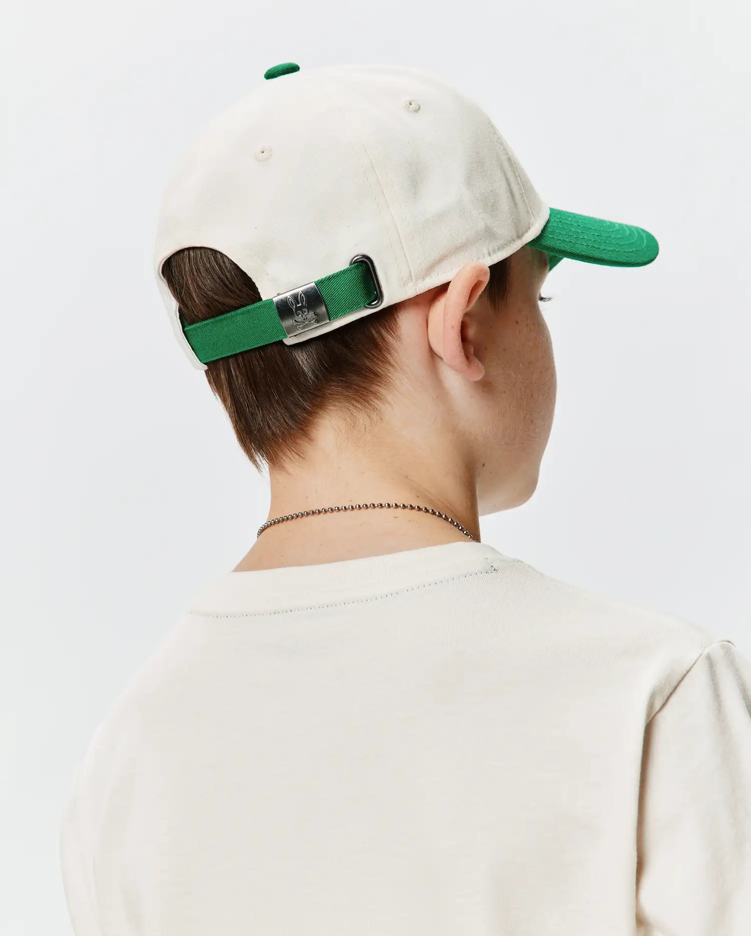 Kids Craig Baseball Cap, an off-white cotton twill cap with a green brim and matching adjustable strap, featuring a silver Psycho Bunny skull logo buckle. Worn with a cream classic fit jersey t-shirt.