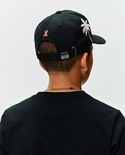 Black Kids Brad Trucker Cap featuring an embroidered orange Psycho Bunny skull logo on the back, a white and pink palm tree graphic on the side, and a metal buckle with an engraved bunny skull.