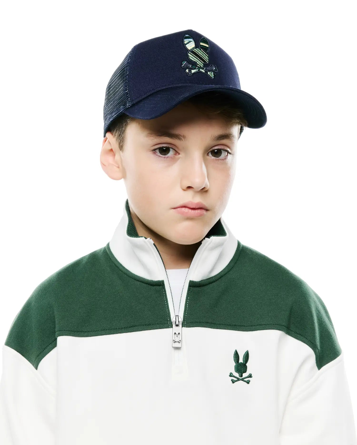 Kids Drew Trucker Cap, navy with a striped green Psycho Bunny logo, and a white and forest green color-block quarter-zip sweatshirt with an embroidered chest logo and branded zipper pull.