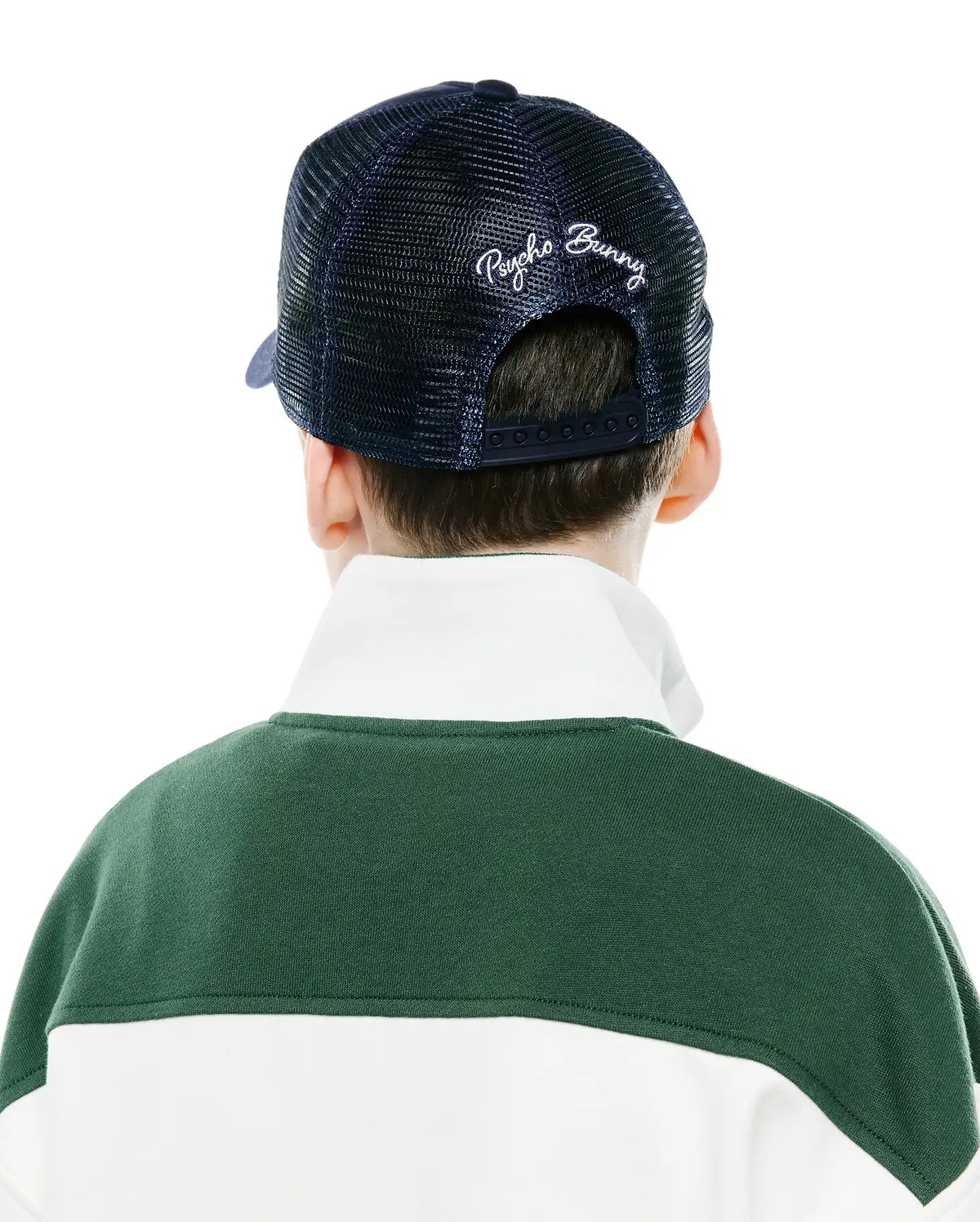 Navy blue mesh Kids Drew Trucker Cap with white embroidered Psycho Bunny script on the back, worn with a dark green and white color-block long-sleeve top featuring a crisp white collar.