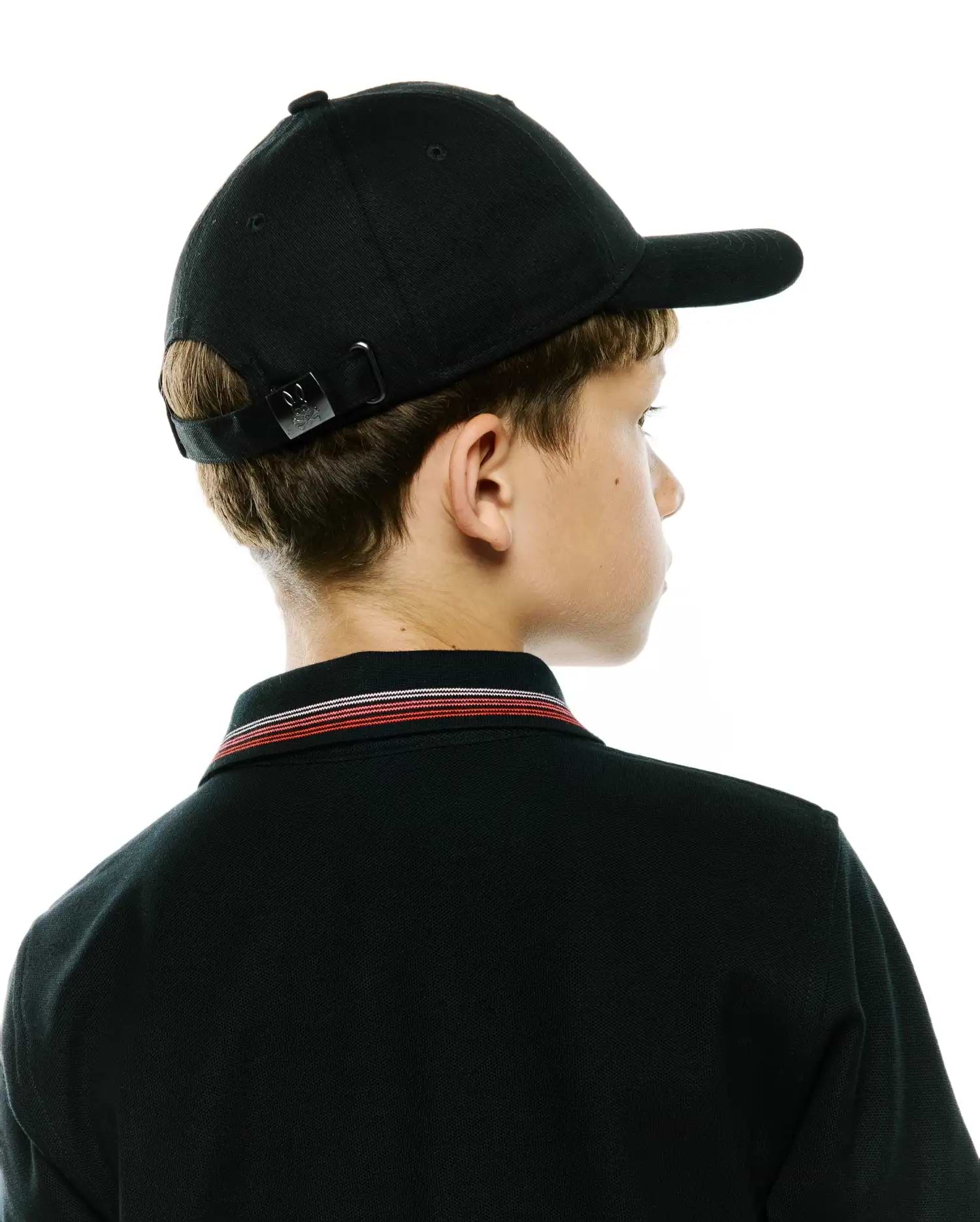 Black pique polo with red, white, and black striped collar, paired with a black Kids Jaydon Baseball Cap featuring a small Psycho Bunny skull logo on its adjustable metal buckle.