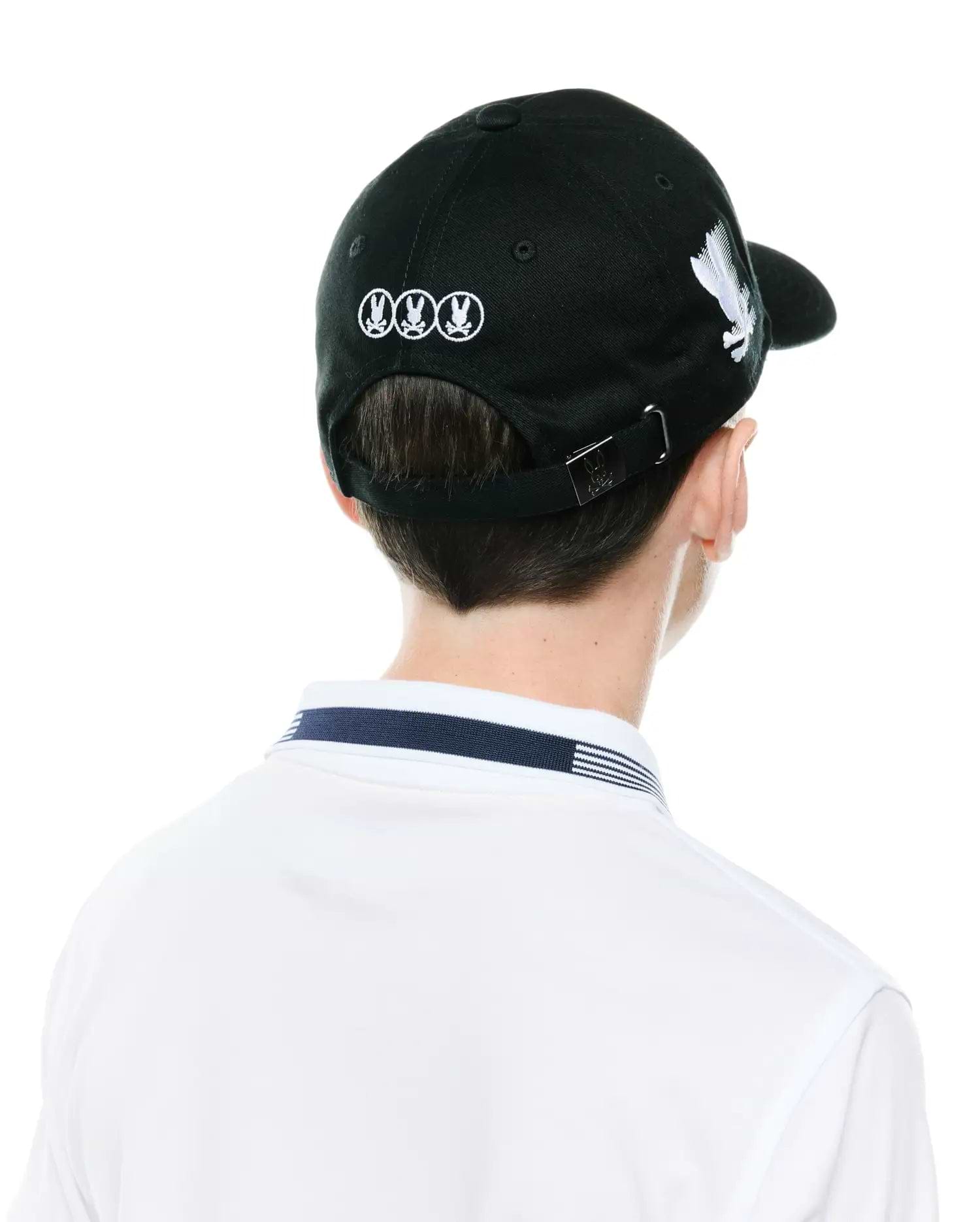 Black Kids Nathaniel A-Frame Trucker Cap with white embroidered Psycho Bunny skull logos on back and side, metal logo buckle, paired with a white polo featuring a navy and white striped collar.