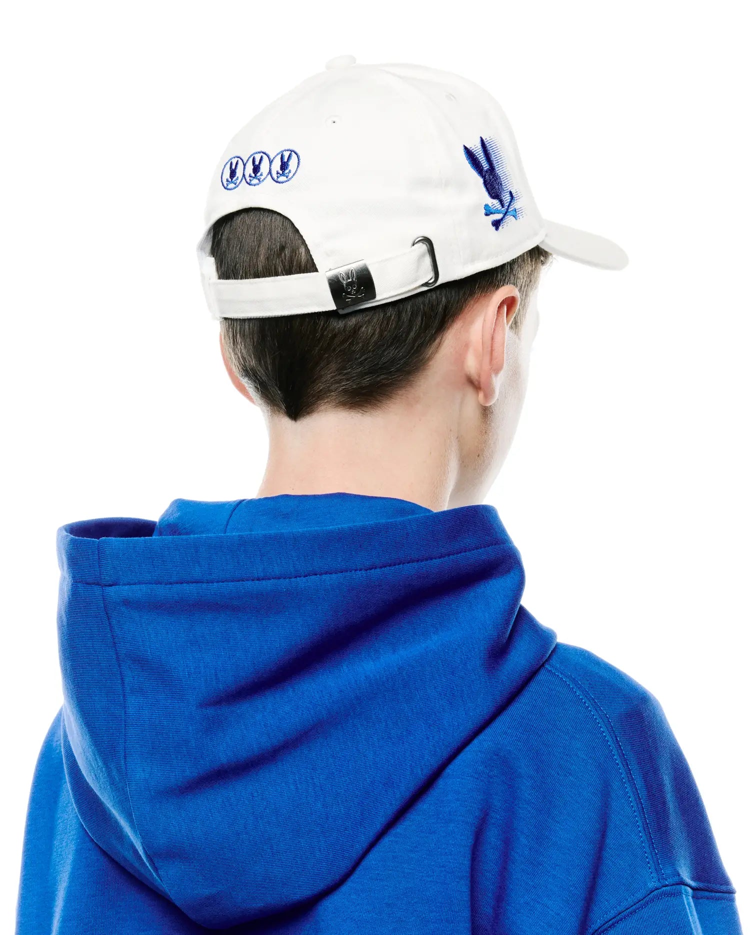 Kids Nathaniel A-Frame Trucker Cap in white cotton twill with blue embroidered Psycho Bunny skull logos on the side and back, metal buckle, and a relaxed royal blue jersey hoodie.