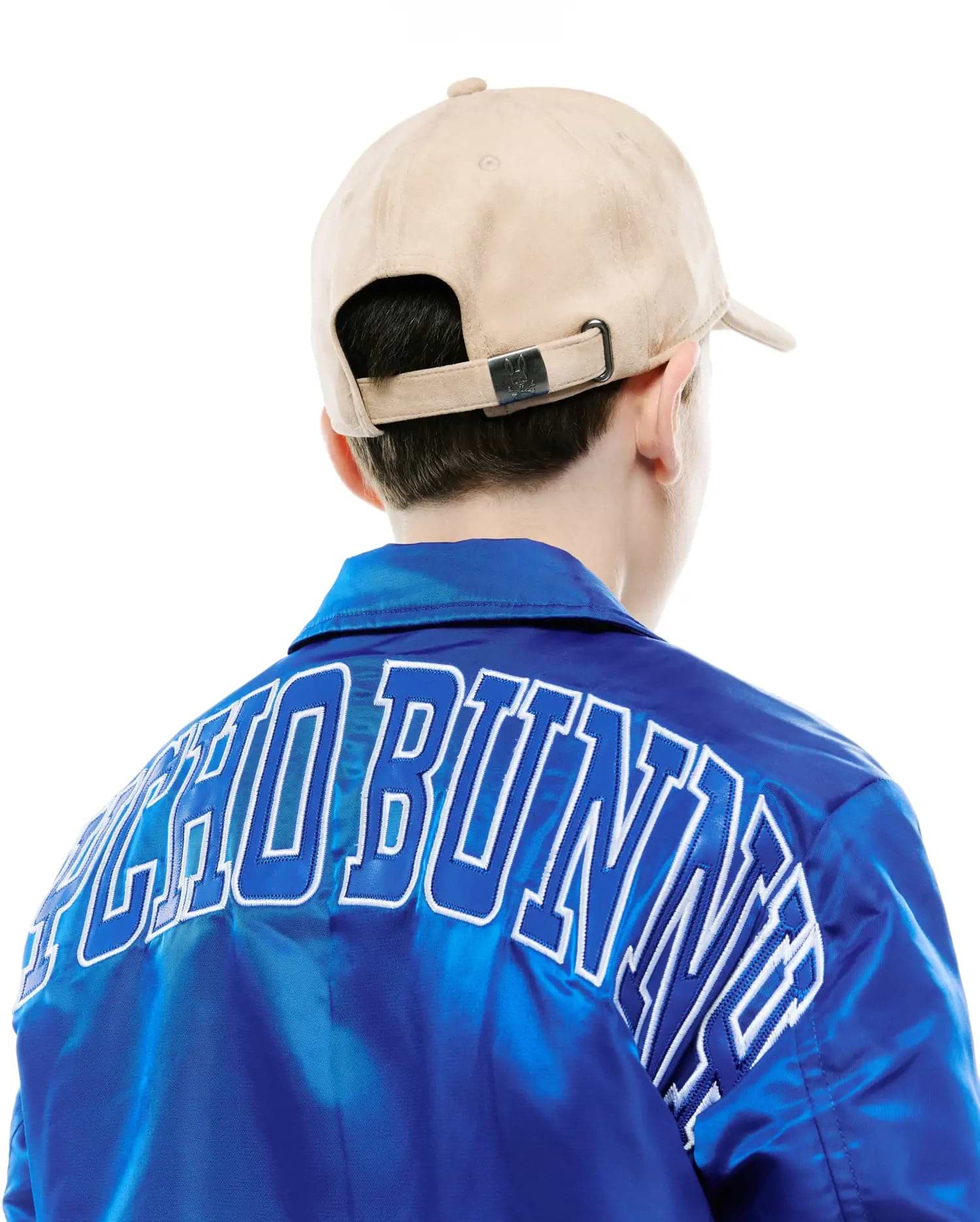 Royal Blue satin jacket with large white-outlined PSYCHO BUNNY embroidery on the back, paired with a beige suede Kids Kenneth A-Frame Trucker Cap featuring a Psycho Bunny logo buckle.