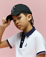 A young person stands against a pink background, confidently tilting the brim of the Psycho Bunny KIDS ARCHIVE BASEBALL CAP (KO1500004), paired with a white polo shirt featuring dark blue trim.