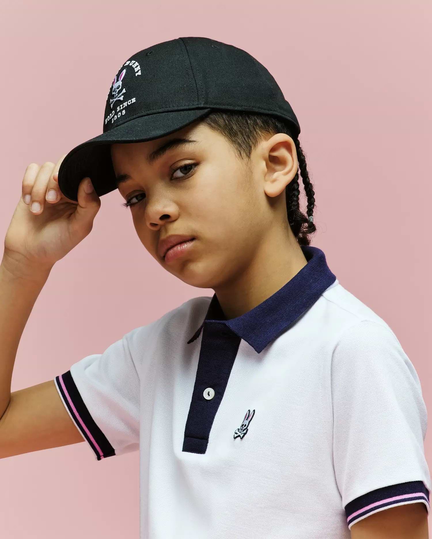 A young person stands against a pink background, confidently tilting the brim of the Psycho Bunny KIDS ARCHIVE BASEBALL CAP (KO1500004), paired with a white polo shirt featuring dark blue trim.