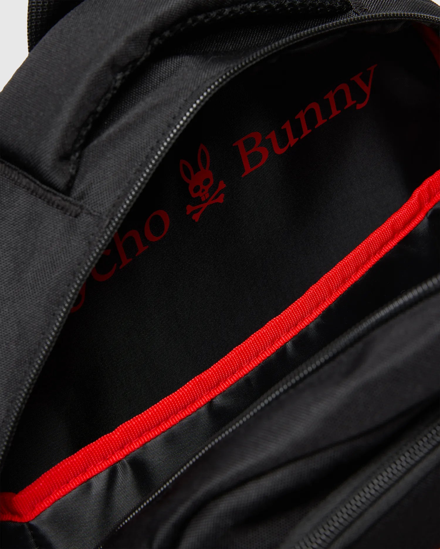 Black Kids Pal Backpack with a durable synthetic exterior. The interior lining is black with a large, bold red Psycho Bunny skull logo and text, accented by a vibrant red webbing trim along the zipper.