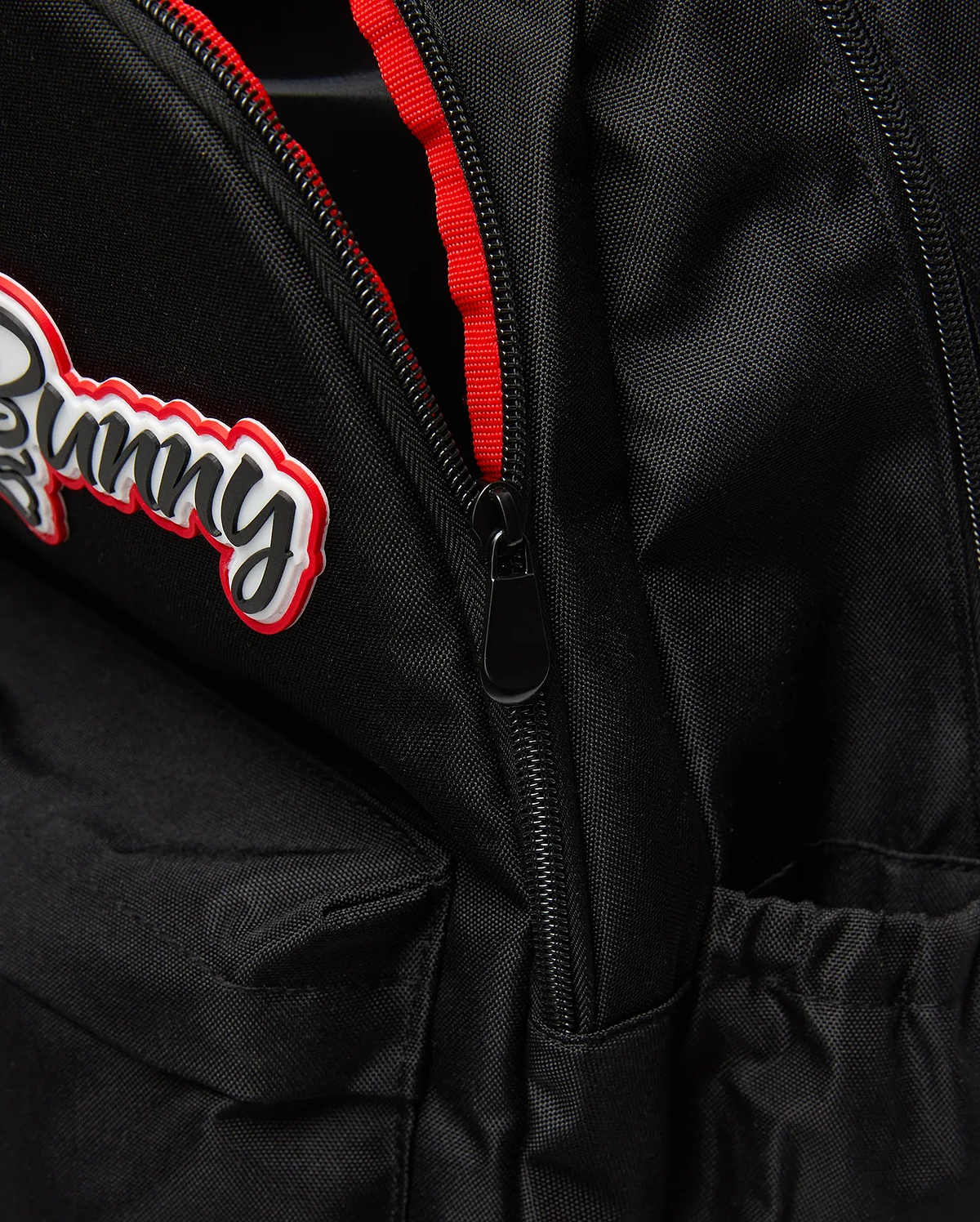 Black Kids Pal Backpack of durable woven fabric, featuring a raised black script Bunny logo with white and red outlines, a black zipper with red lining, and an elasticated side pocket.