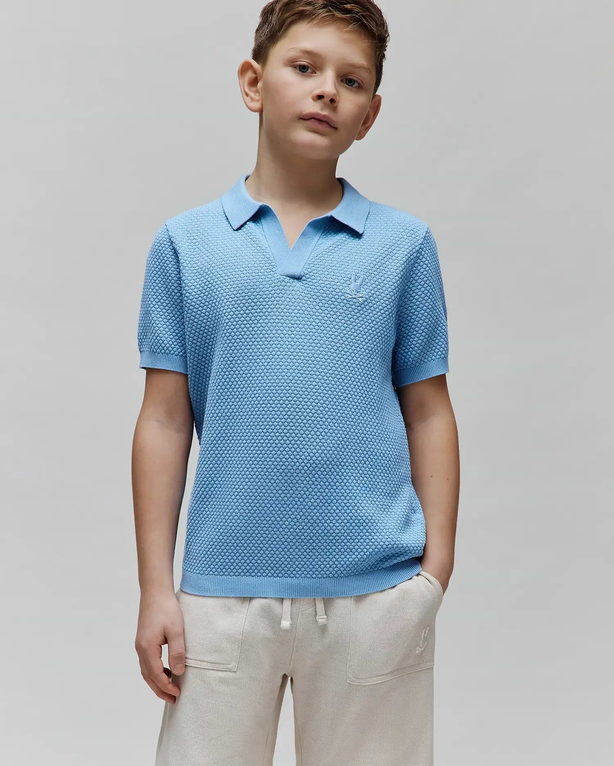 Kids Devon Bubble Stitch Knit Polo in light blue, a classic fit with an open V-neck collar, ribbed cuffs, and a small embroidered Psycho Bunny skull logo on the left chest.