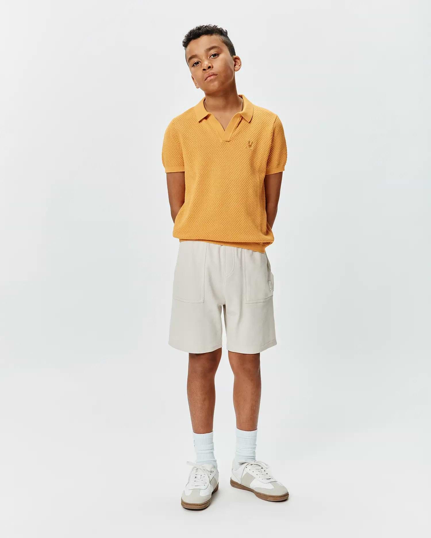 Kids Devon Knit Polo in mustard yellow, featuring a textured knit fabric, V-neck, and small embroidered Psycho Bunny skull logo on the left chest, paired with relaxed fit light beige shorts with patch pockets.
