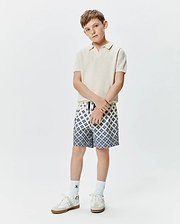 Kids Devon Knit Polo in cream, featuring a textured knit fabric and small embroidered Psycho Bunny logo on the chest, paired with blue and white geometric pattern shorts.