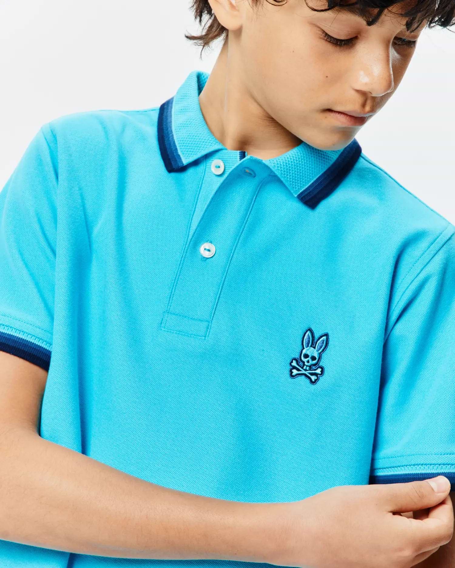 Kids Holden Pique Polo in vibrant aqua blue, classic fit, textured pique fabric, two-button placket, and ribbed collar and cuffs with navy and light blue stripes. Small embroidered Psycho Bunny skull logo on left chest.