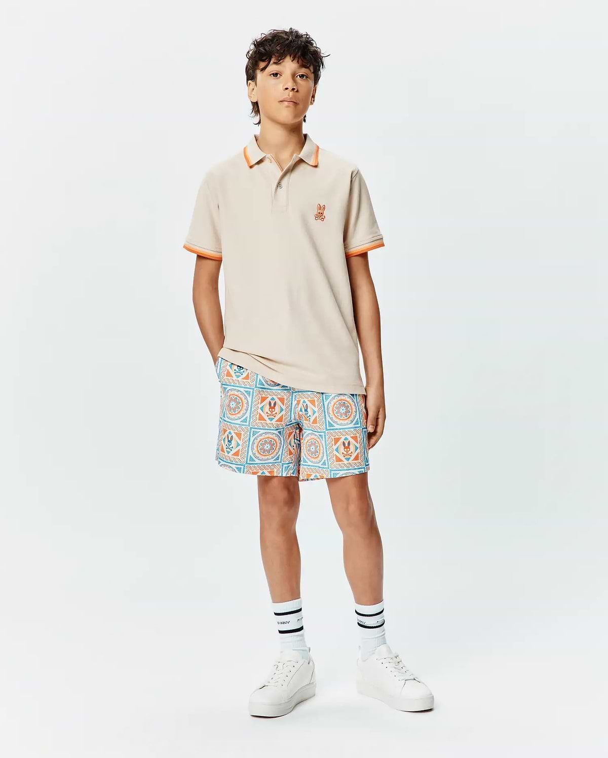 Kids Holden Pique Polo in beige with orange tipped collar and cuffs, featuring a small embroidered orange Psycho Bunny skull logo on the left chest, paired with patterned shorts.