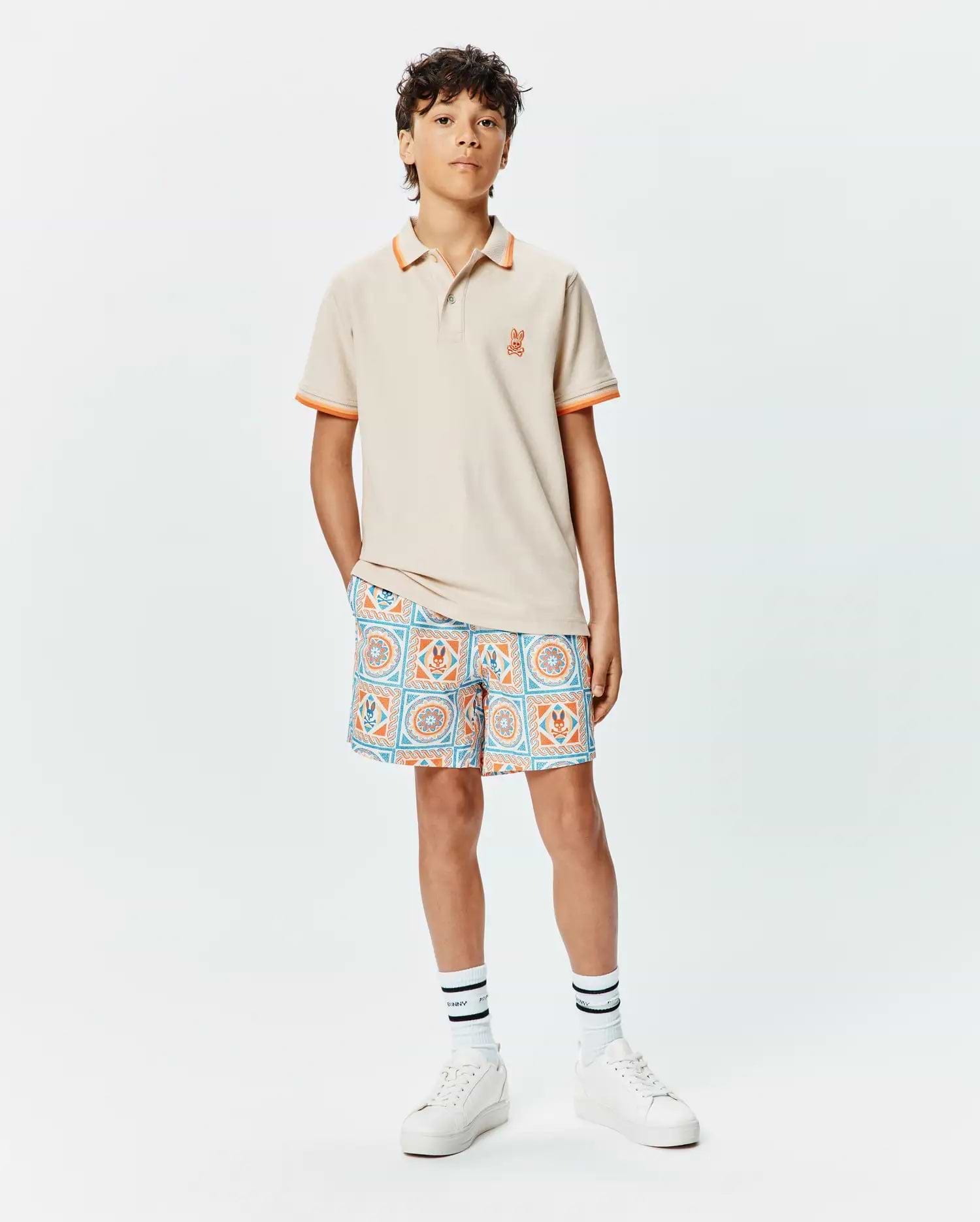Kids Holden Pique Polo in beige with orange tipped collar and cuffs, featuring a small embroidered orange Psycho Bunny skull logo on the left chest, paired with patterned shorts.