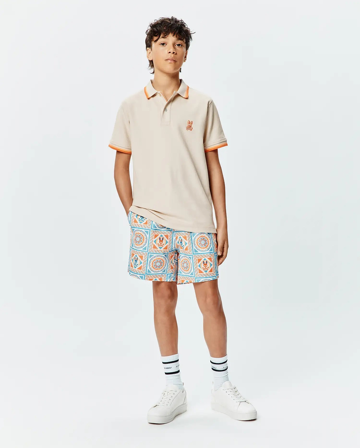 Kids Holden Pique Polo in beige with orange tipped collar and cuffs, featuring a small embroidered orange Psycho Bunny skull logo on the left chest, paired with patterned shorts.