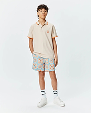 Kids Holden Pique Polo in beige with orange tipped collar and cuffs, featuring a small embroidered orange Psycho Bunny skull logo on the left chest, paired with patterned shorts.