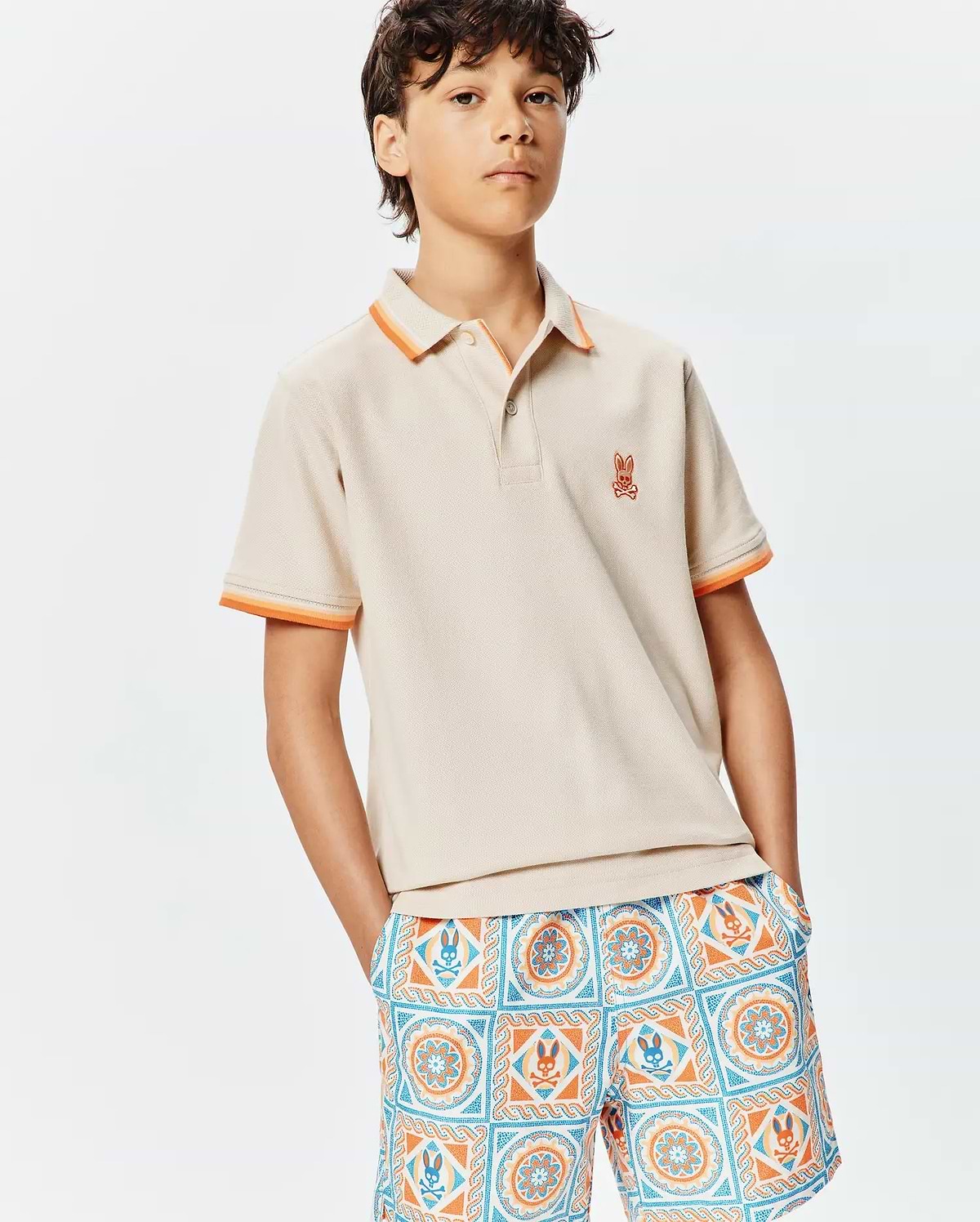 Kids Holden Pique Polo in beige, classic fit, with orange and light orange striped pique collar and cuffs, featuring a small embroidered orange Psycho Bunny skull logo on the left chest.