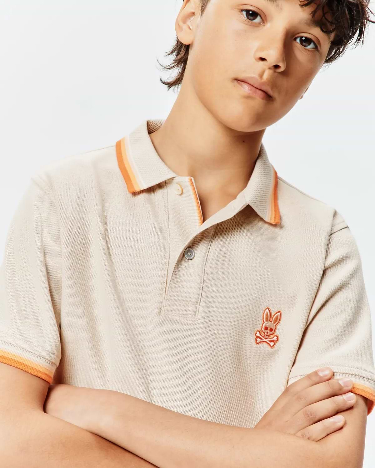 Kids Holden Pique Polo in beige pique, featuring a classic fit, orange striped collar and cuffs, with a small embroidered orange Psycho Bunny skull logo on the left chest.