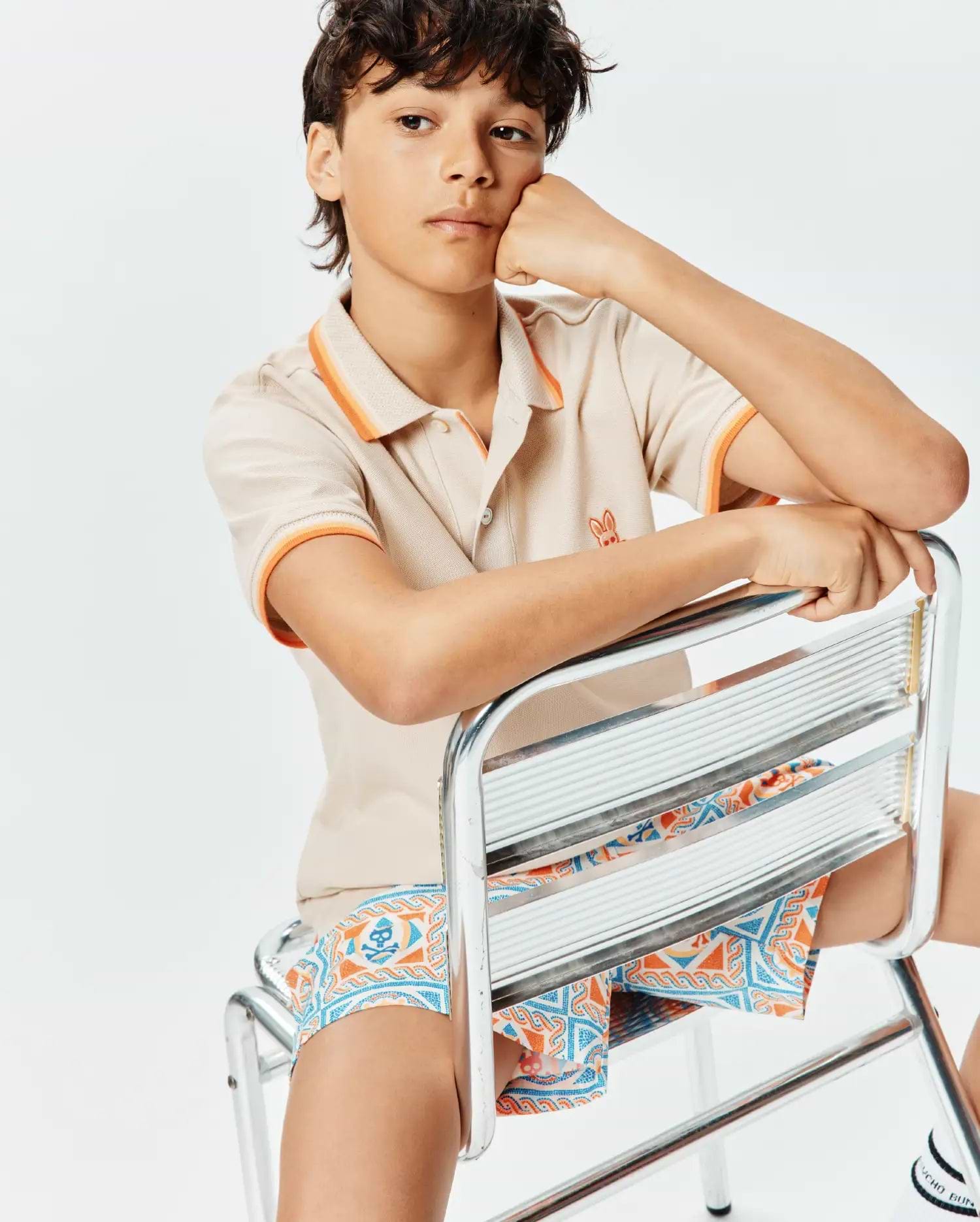 Kids Holden Pique Polo in beige pique with orange tipping on collar and cuffs, featuring a small embroidered orange Psycho Bunny logo on the chest, paired with blue and orange patterned shorts with skull motifs.