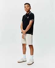 Kids Brad Pique Polo in black with white collar and sleeve tipping, featuring an embroidered Psycho Bunny skull logo on the left chest, paired with light beige knit shorts with an embroidered Psycho Bunny logo. (4)
