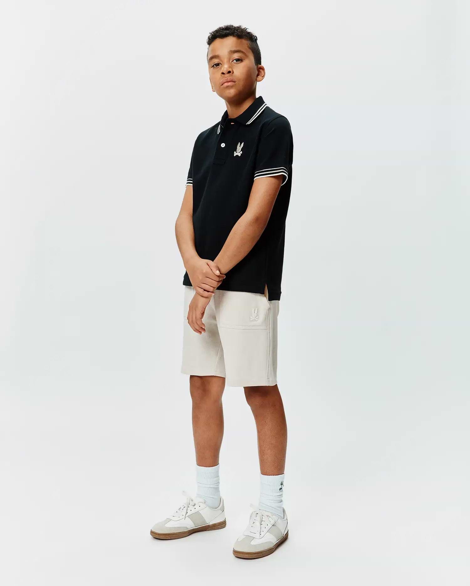 Kids Brad Pique Polo in black with white collar and sleeve tipping, featuring an embroidered Psycho Bunny skull logo on the left chest, paired with light beige knit shorts with an embroidered Psycho Bunny logo. (4)