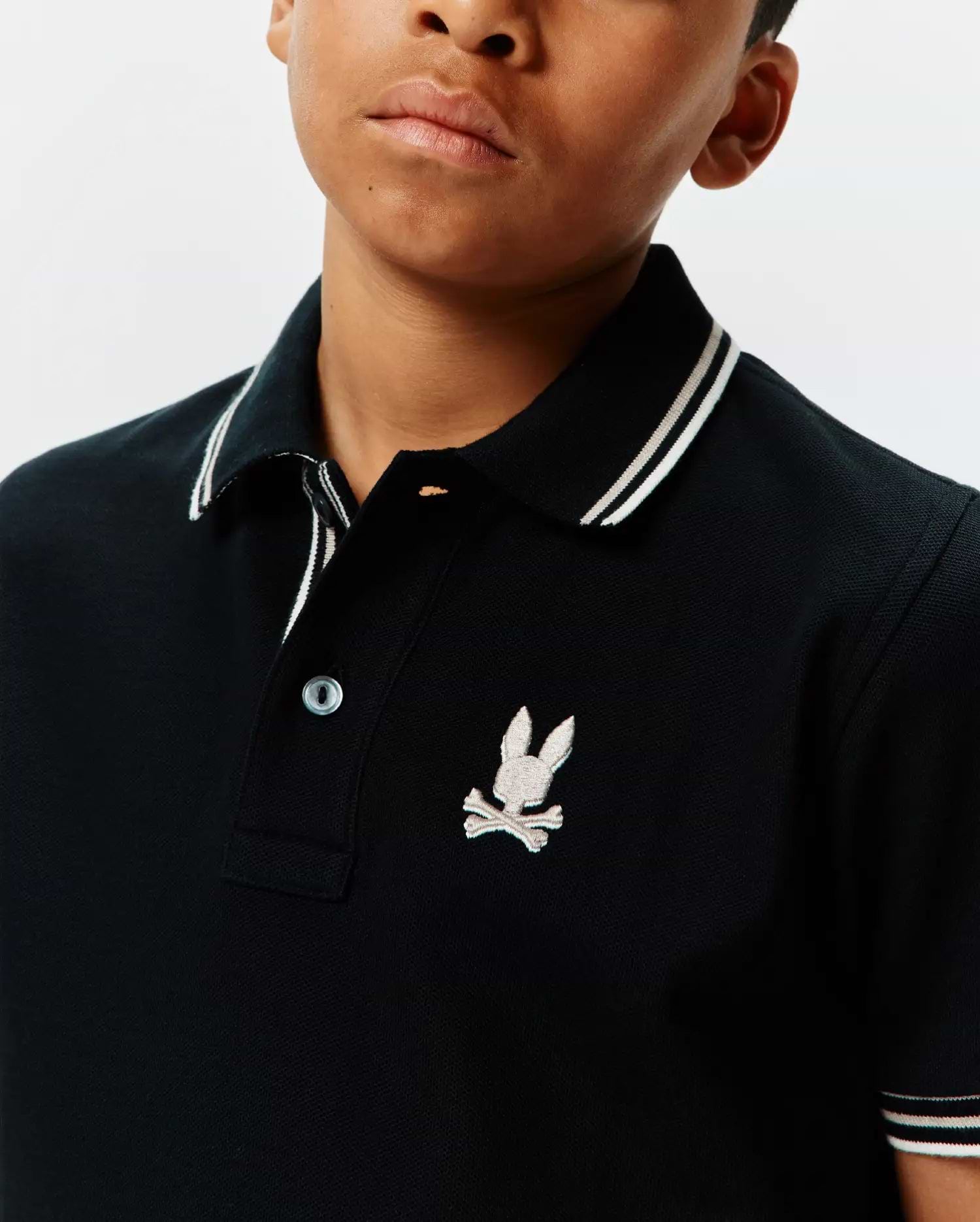 Kids Brad Pique Polo, black classic pique polo with white striped collar and sleeve cuffs, featuring an embroidered light grey Psycho Bunny skull logo on the left chest. (3)