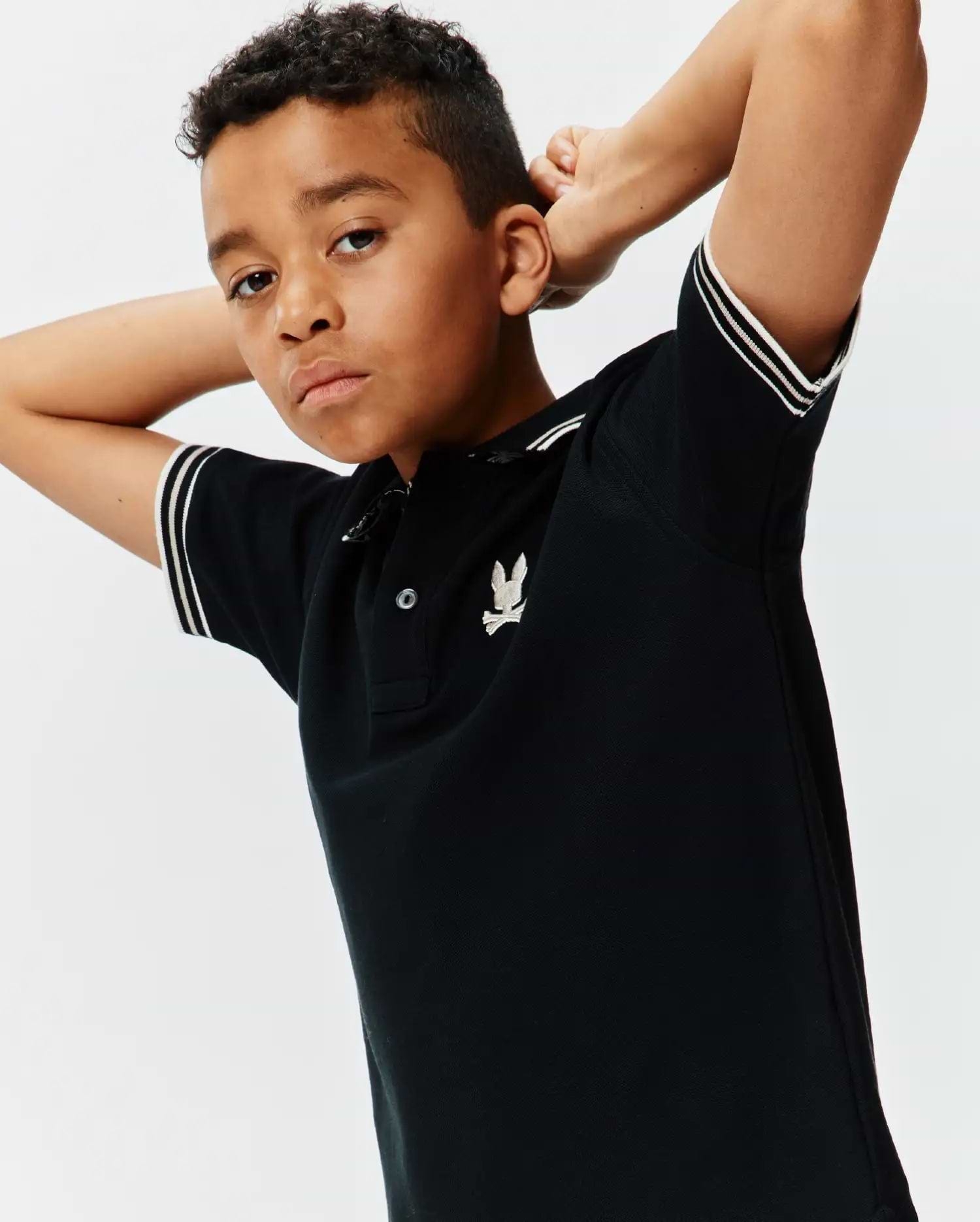 Kids Brad Pique Polo, a classic black pique polo shirt with a two-button placket, ribbed collar, and double white striped sleeve cuffs, featuring a small embroidered silver Psycho Bunny skull logo on the left chest. (1)