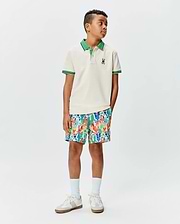 Kids Sunny Pique Polo, a classic fit cream pique polo with green collar, striped cuffs, and small embroidered Psycho Bunny skull logo, paired with vibrant multi-color bunny print shorts.