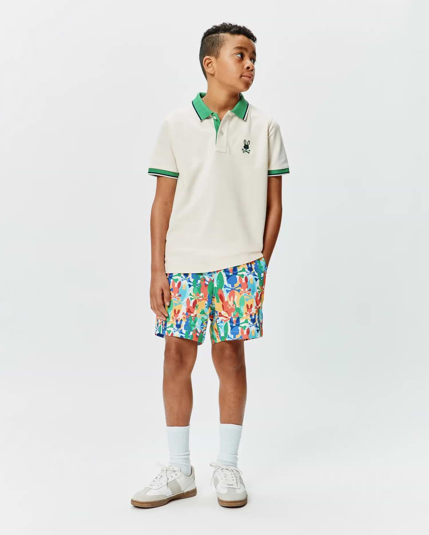 Kids Sunny Pique Polo, a classic fit cream pique polo with green collar, striped cuffs, and small embroidered Psycho Bunny skull logo, paired with vibrant multi-color bunny print shorts.