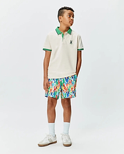 Kids Sunny Pique Polo, a classic fit cream pique polo with green collar, striped cuffs, and small embroidered Psycho Bunny skull logo, paired with vibrant multi-color bunny print shorts.