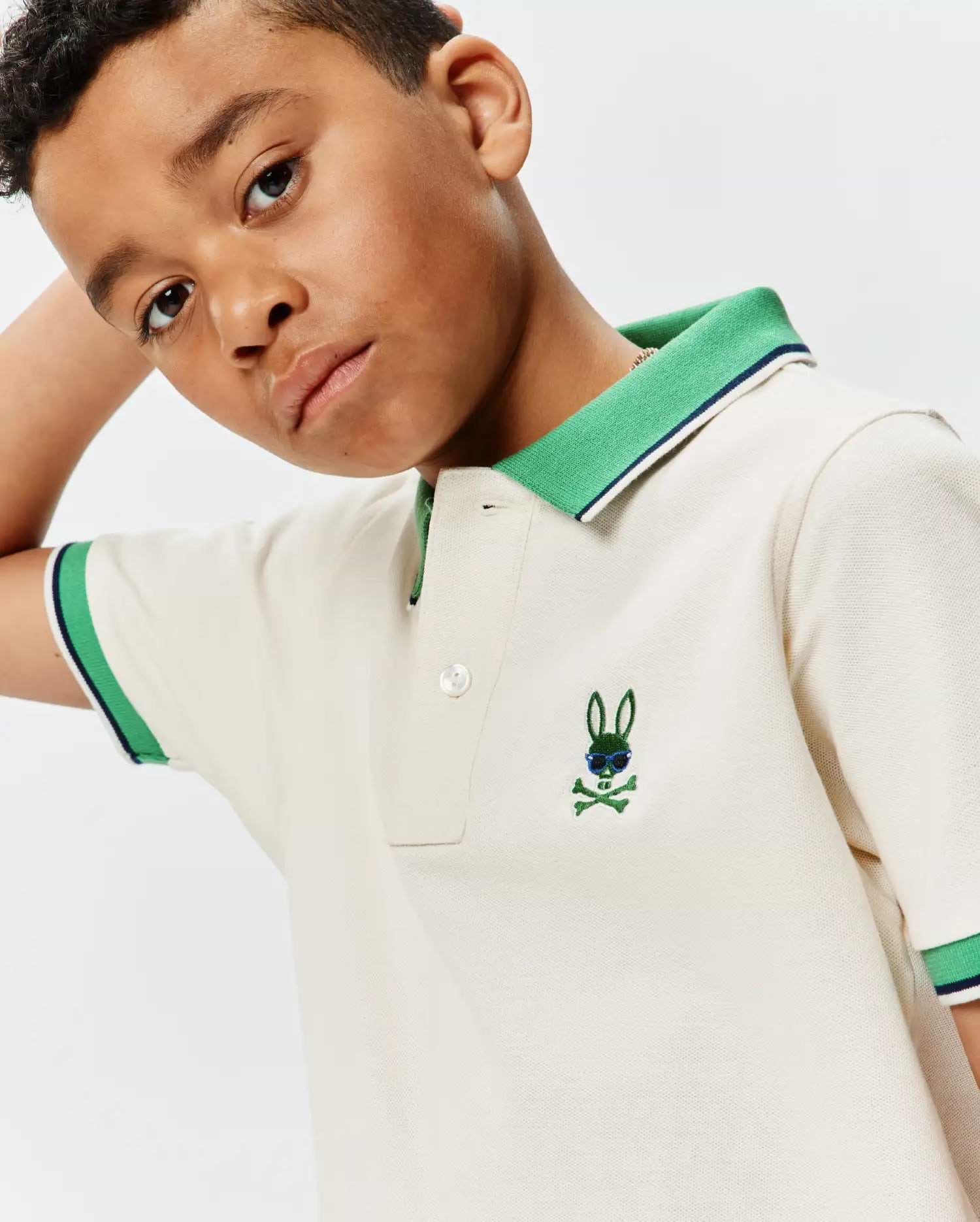 Kids Sunny Pique Polo in cream, featuring a classic fit, green ribbed collar with navy stripe, matching striped cuffs, and a small embroidered green Psycho Bunny skull with blue sunglasses on the left chest.