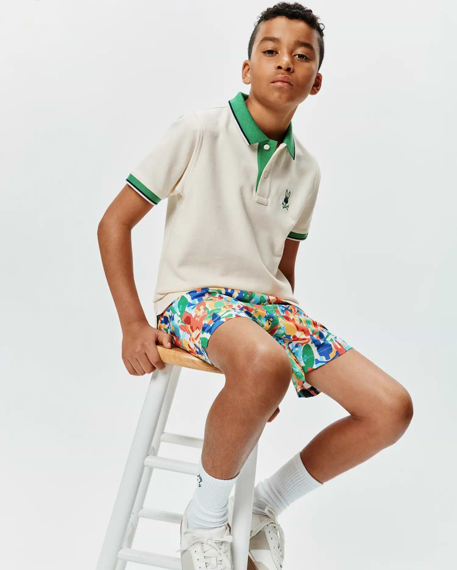 Kids Sunny Pique Polo in cream pique with a green collar and striped cuffs, featuring a small embroidered Psycho Bunny logo, paired with multi-color abstract print shorts.