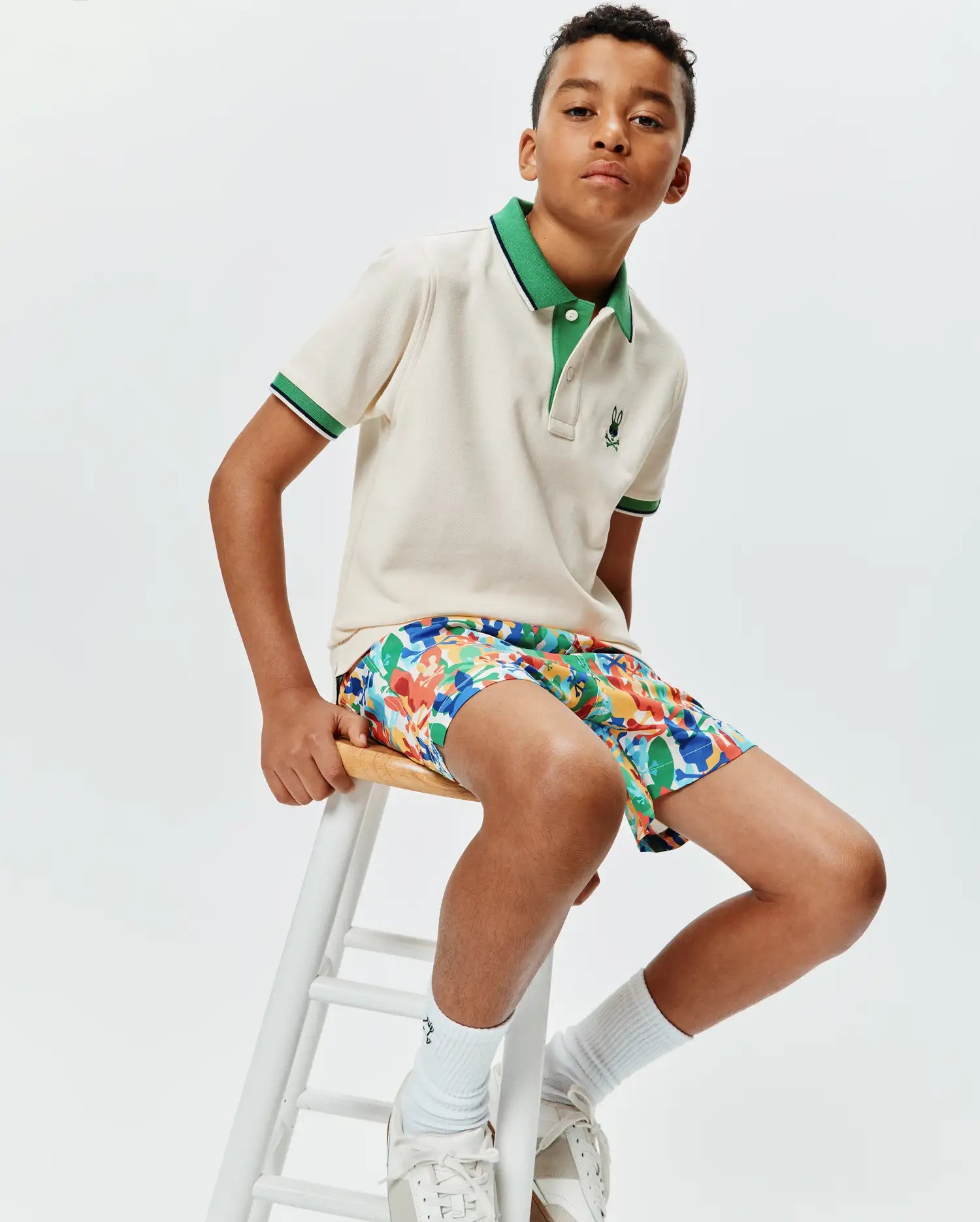 Kids Sunny Pique Polo in cream pique with a green collar and striped cuffs, featuring a small embroidered Psycho Bunny logo, paired with multi-color abstract print shorts.