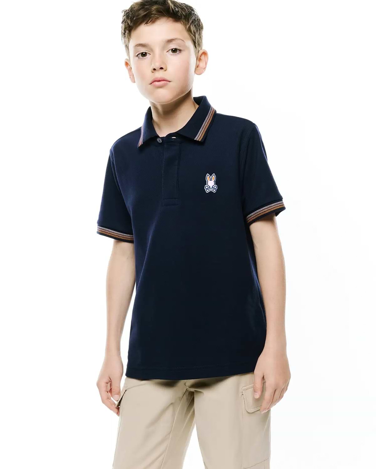 Kids Jaydon Pique Polo, a navy classic fit pique polo with orange, white, and brown striped collar and sleeve cuffs, featuring an embroidered Psycho Bunny skull logo on the left chest.