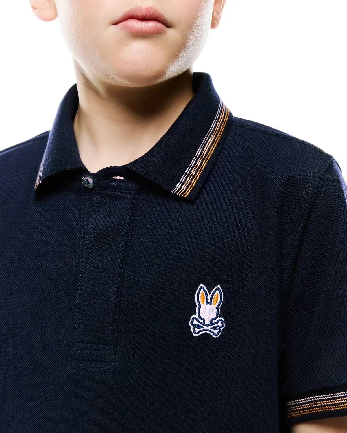 Kids Jaydon Pique Polo in navy, classic fit, pique texture. Features small embroidered multi-color Psycho Bunny skull logo on left chest, with orange and white striped collar and cuffs.
