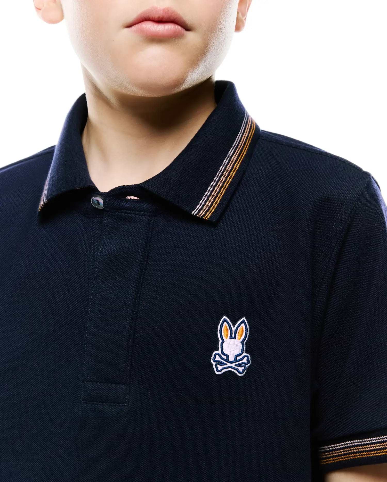 Kids Jaydon Pique Polo in navy, classic fit, pique texture. Features small embroidered multi-color Psycho Bunny skull logo on left chest, with orange and white striped collar and cuffs.