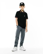 KIDS JAYDON PIQUE POLO, a black classic fit pique polo with red/white striped collar/cuffs, featuring a small embroidered Psycho Bunny logo. Worn with grey pants, white sneakers, and a black cap with a large Psycho Bunny logo.
