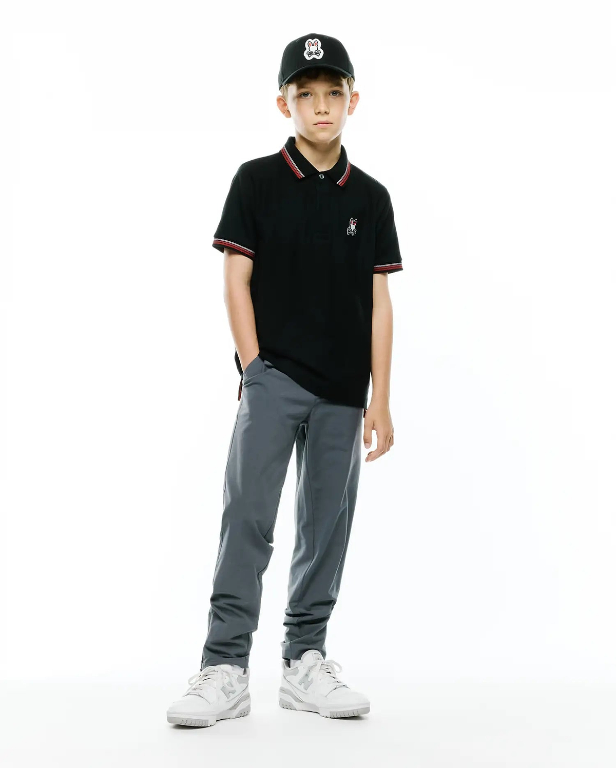 KIDS JAYDON PIQUE POLO, a black classic fit pique polo with red/white striped collar/cuffs, featuring a small embroidered Psycho Bunny logo. Worn with grey pants, white sneakers, and a black cap with a large Psycho Bunny logo.