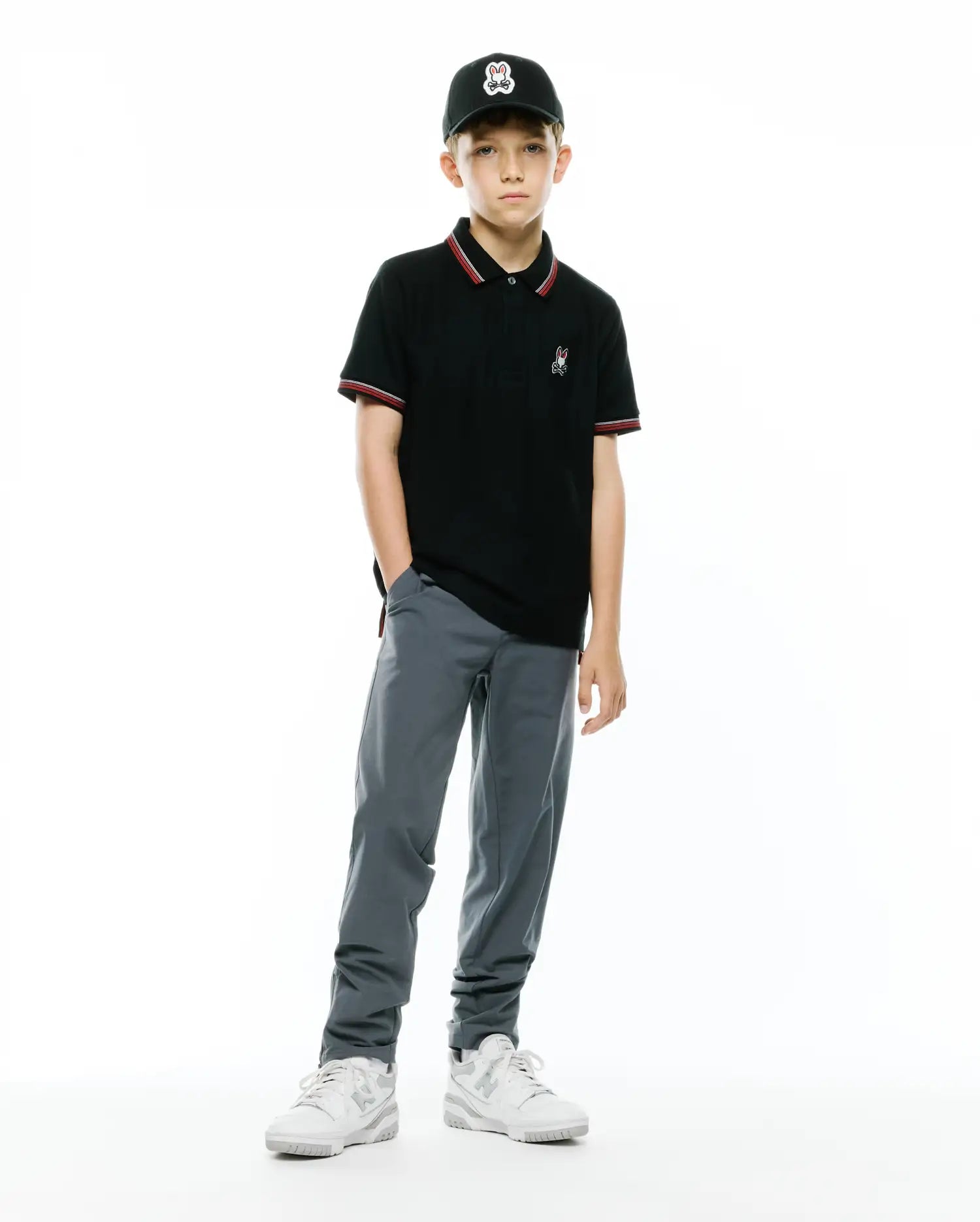 KIDS JAYDON PIQUE POLO, a black classic fit pique polo with red/white striped collar/cuffs, featuring a small embroidered Psycho Bunny logo. Worn with grey pants, white sneakers, and a black cap with a large Psycho Bunny logo.