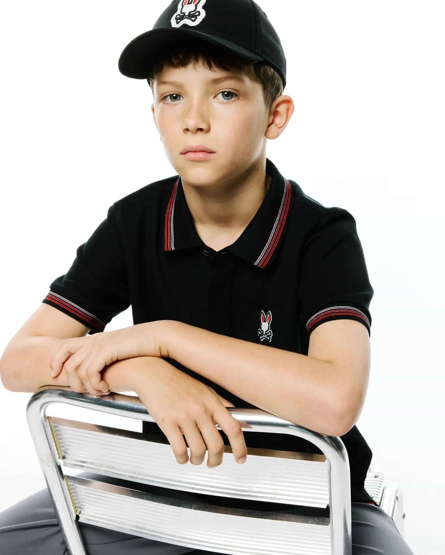 Kids Jaydon Pique Polo in black, with red and white striped collar and cuffs, a small embroidered Psycho Bunny skull logo on the left chest. Also, a black baseball cap with a large embroidered bunny skull logo.