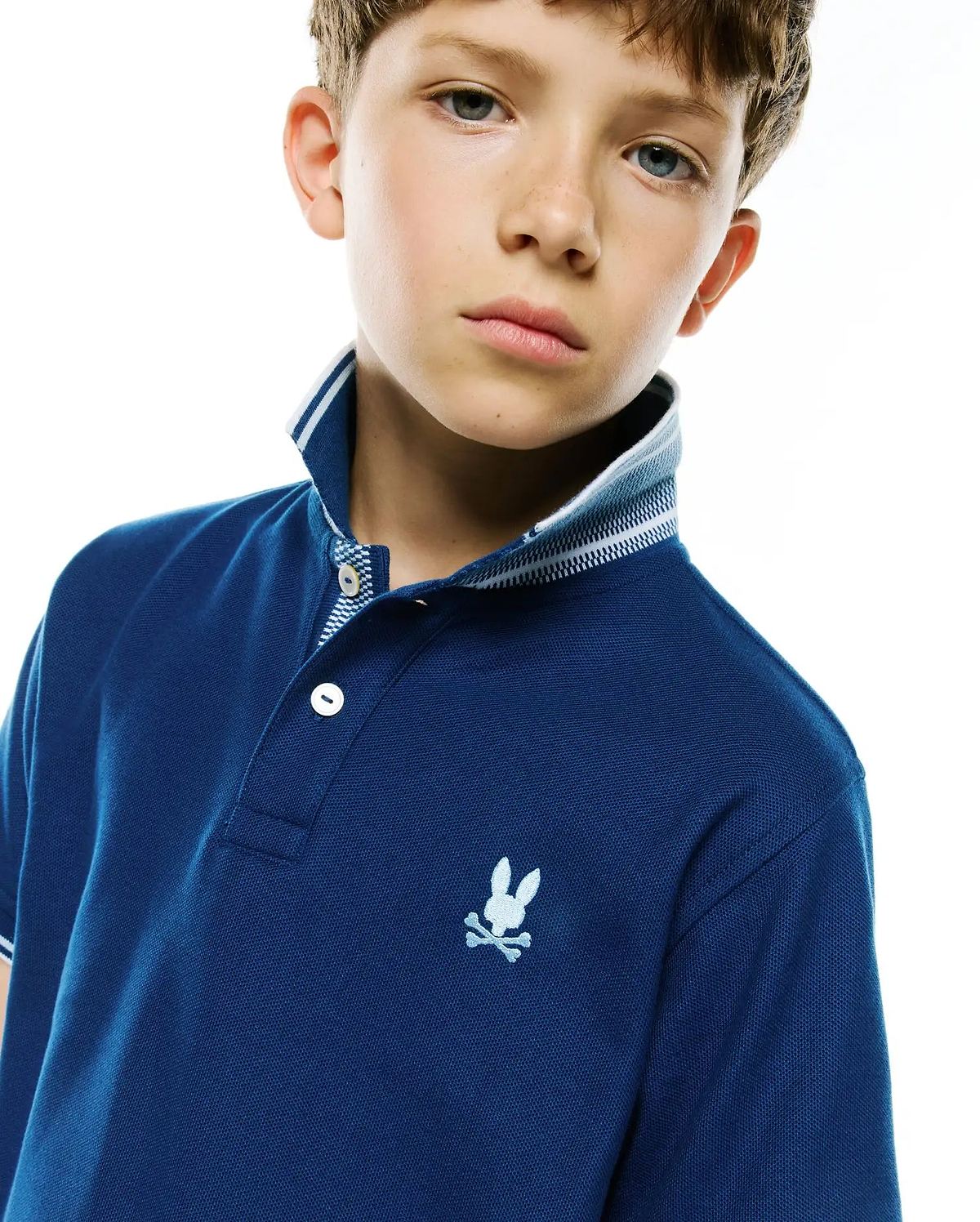 Kids Stan Pique Polo, deep blue pique polo shirt with white double-striped ribbed collar, three-button placket with patterned lining, and small embroidered white Psycho Bunny skull logo on left chest.